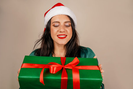 Studio shot of beautiful happy joyful excited girl in santa hat and green dress holding gift box in...