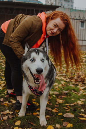 A young girl with long red hair plays with a husky dog. A woman walks her dog in the park.の写真素材