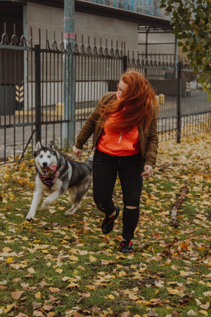 A young girl with long red hair plays with a husky dog. A woman walks her dog in the park.の写真素材