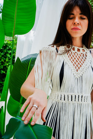 A young brunette woman in a white macrame cape stands gracefully against a lush green garden. A...