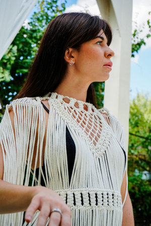 A young brunette woman in a white macrame cape stands gracefully against a lush green garden. A...