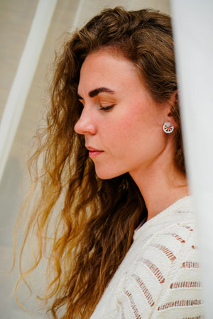 A close-up of a blond woman against a white tulle background, dressed in a delicate white knitted outfit. The girl has an earring in her ear, adding sparkle.の写真素材