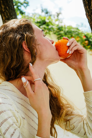 A young woman with curly hair holds a ripe peach, enjoying its aroma. She is dressed in white and accessorized with silver rings, exuding natural elegance.の写真素材