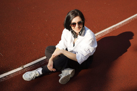 Young woman in casual outfit and headphones sits on a track in the sunlight, enjoying music and...