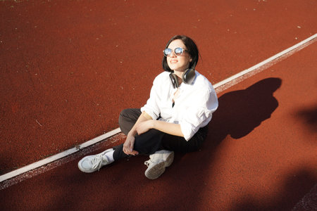 Young woman in casual outfit and headphones sits on a track in the sunlight, enjoying music and relaxation outdoors.の写真素材