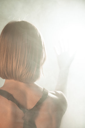A woman in an apron poses with her back to the camera, creating an artistic and minimalist composition. In contrasting light, the woman poses in a light smoke.の写真素材