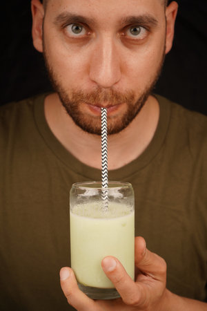 Close-up of a man drinking green matcha latte from a glass through a straw, tea culture concept. A man sips tea through a straw.の写真素材