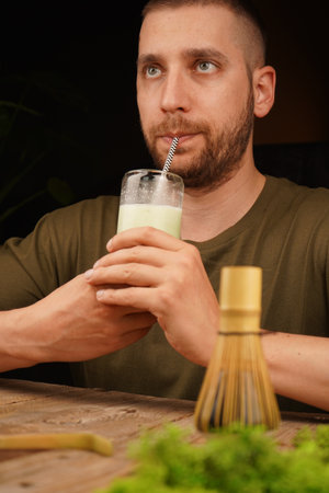 A thoughtful man enjoys a glass of creamy matcha latte with a bamboo whisk on a wooden table. Ideal for themes of mindfulness, wellness, and tea culture.の写真素材