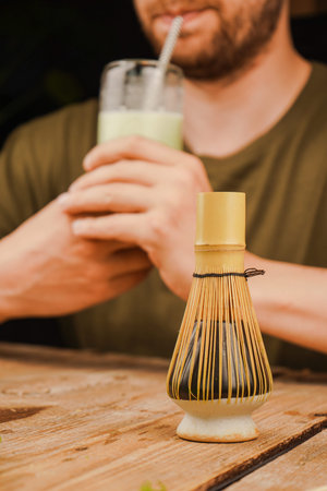 A thoughtful man enjoys a glass of creamy matcha latte with a bamboo whisk on a wooden table. Ideal...