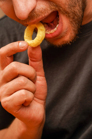 A close-up of a man enjoying a crunchy snack in the shape of a ring. A bearded man holds a golden snack to his mouth, emphasizing its crunch.の写真素材