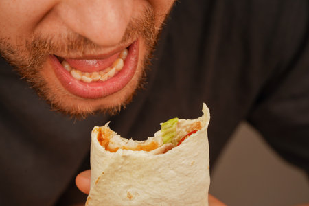 A man opens his mouth to take a bite of a lavash wrap filled with meat, salad, and fresh veggies. His beard adds character to the dynamic scene.の写真素材