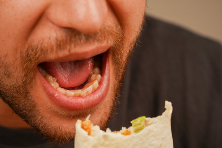 Close-up of a bearded man holding and eating a meat roll with fresh vegetables. Captures the anticipation of the first bite.の写真素材