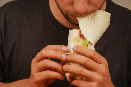 Close-up of a bearded man holding and eating a meat roll with fresh vegetables. Captures the anticipation of the first bite.の写真素材