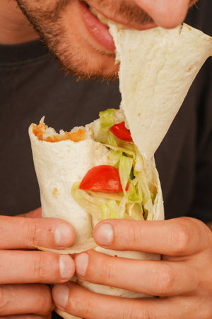 A man with a small beard bites into a pita roll filled with meat, fresh vegetables, and salad. The photo, taken in mid-motion, emphasizes the delicious food and its enjoyment.の写真素材