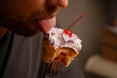 A young man in a dark T-shirt licks a cupcake skewered on a fork. A man enthusiastically eats a...