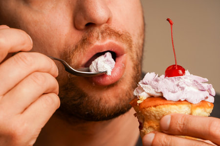 Close-up of a man enjoying a piece of cake with a cherry and cream frosting with a spoon. As a concept, life in pleasure, dessert and the desire for something sweet.の写真素材