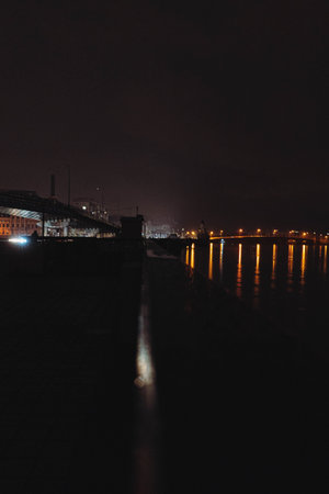 Side view of a colorful bridge illuminated by white lights at night. The bridge stands on the river. Yellow light is reflected in the water.の写真素材