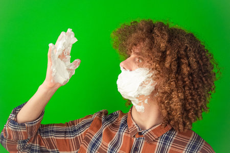 Dressed in a plaid shirt, the curly-haired boy massages shaving foam onto his face, his relaxed...