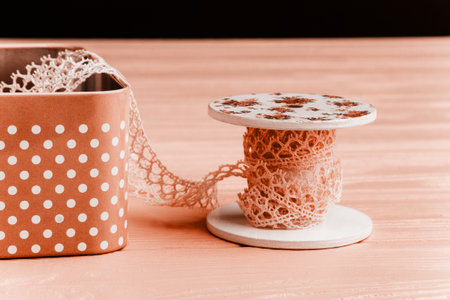 A soft pink lace ribbon on a floral spool alongside a pink polka dot iron box is perfect for crafting and decorating on a soft pink background.の写真素材