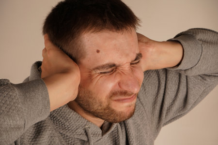Portrait of young man closing his ears by hands and keeping his eyes closed while isolated on grey. Photo of bearded man keeps hands on head and closes eyes.の写真素材