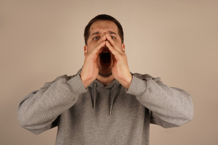 His animated gesture implies shouting or announcing something important. His expression radiates urgency. The dark-haired man holds both palms around his mouth, forming a makeshift megaphone.の写真素材