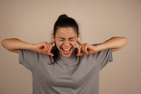 A universal symbol of salvation from auditory overload. Her gesture illustrates a strong need to disconnect from external interference. A woman covers her ears tightly with her fingers, isolating herself from noise.の写真素材