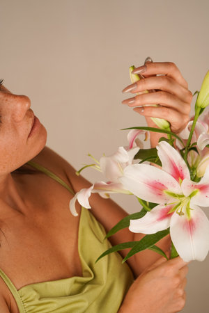 The neutral setting accentuates her sophisticated charm. A young woman in front of a beige wall holds a bouquet of pink lilies, smiling tenderly. Her gaze is full of warmth and care.の写真素材