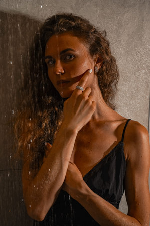 A pensive image of a woman in the shower. A young woman with long hair stands against a gray tiled...
