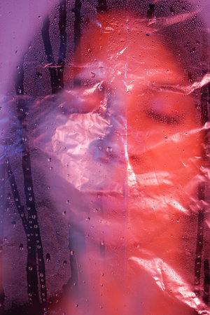 Young adult woman looking at camera with sad expression through plastic wrap. Artistic portrait...