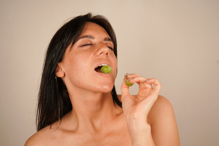 With a playful gaze, the girl bites into a grape, her skin glowing under the soft light, symbolizing freshness and the essence of natural skincare.の写真素材