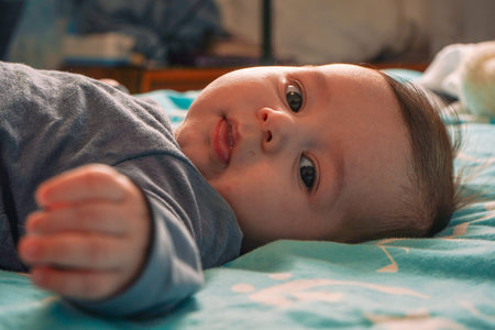 Adorable happy boy in bodysuit lying on back on mattress in crib in home room. View from above. The...