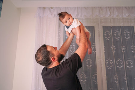 The baby stretches its tiny arms as the father playfully raises them above his head, creating a...