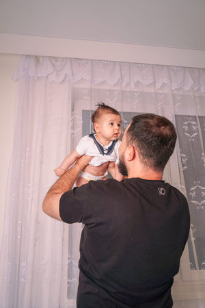 A proud father smiles warmly as he holds his baby high in the air, celebrating the joys of...