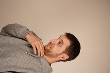Neutral background highlights the simplicity of the scene. Young man in a gray hoodie standing indoors. He appears to be engaged in thought or mid-gesture, creating a natural and casual moment.の写真素材