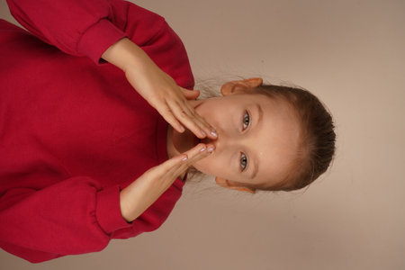 With hands near her mouth, the girl pretends to shape a smile. Her playful expression radiates happiness and creativity. A delightful gesture capturing her carefree mood.の写真素材