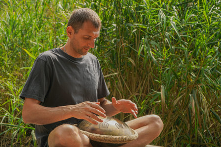 Seated in the lotus position by a calm river, a man plays a handpan, combining serene melodies with...