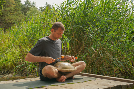 A young man sits in the lotus position on a wooden pier by the river. The guy plays a metal handpan...