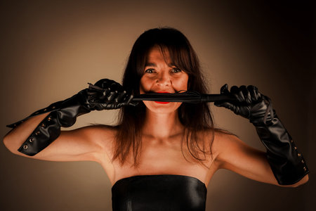 A striking woman with long dark hair bites a piece of a black leather whip, wearing elbow-length...