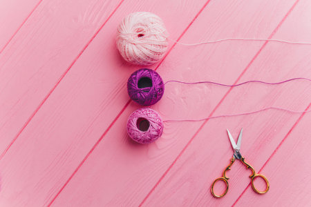 A roll of pink and purple skeins of thread and iron scissors lie on a wooden pink surface, perfect...