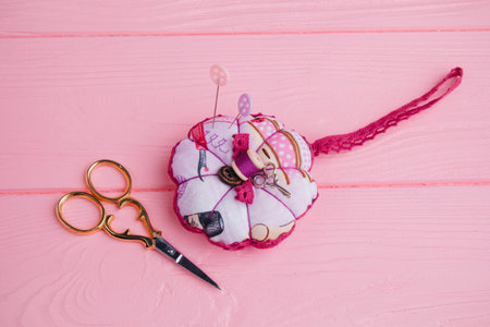 A pincushion rests on pink boards. Scissors for needlework lie next to needles stuck in a sectional...