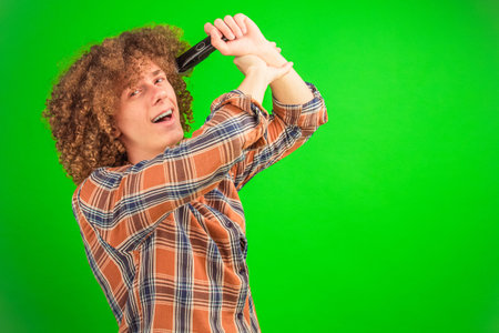 The boy with unruly curls wears a casual shirt while shaving his hair with a trimmer, the solid...