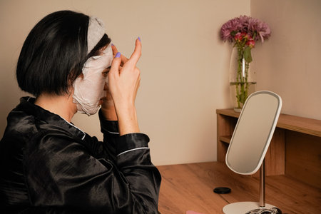 A small mirror reflects her calm expression. Sitting at a makeup table, a woman relaxes with a hydrating sheet mask. Black pajamas and a neat headband complete the scene.の写真素材