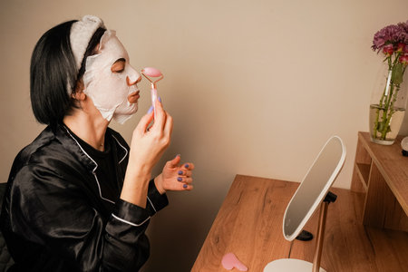 A small mirror adds charm to the scene. A young woman is using a hydrating mask at her dressing table. She is wearing elegant black pajamas and a headband.の写真素材