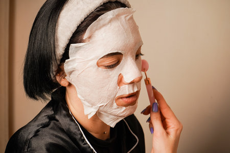 A woman uses a roller massager on her neck. Beauty treatments at home. Sitting at a beauty table, a woman uses a sheet mask to moisturize her skin. Black silk pajamas add a touch of luxury.の写真素材