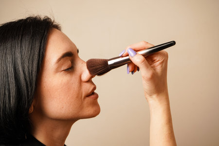 With a steady hand, a woman uses a brush to apply makeup to her face. Her smooth skin and natural...