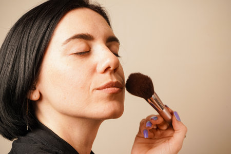 A close-up of a woman applying makeup captures her gentle technique with a fluffy brush. Her closed eyes convey concentration. The minimalist background enhances the focus on her routine.の写真素材