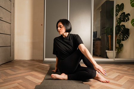 Sitting on a yoga mat, a girl in black leggings and a sports t-shirt performs stretches in her cozy...