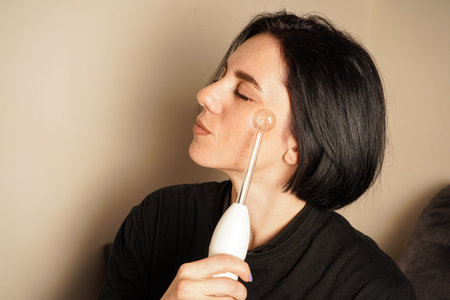 A woman is taking care of her face with a white high-frequency darsonval. Her focused expression and...