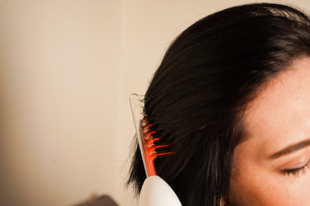 Her calm demeanor and simple background emphasize the importance of darsonvalization. Perfect for health, skin care, and wellness topics.Sitting indoors, a woman applies a beauty gadget to her scalp.の写真素材