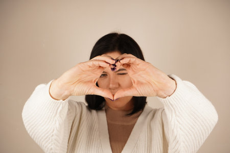 Smiling gently, the woman makes a heart gesture with her hands, symbolizing care and connection. Her relaxed posture conveys calm and sincerity. The gesture forms a heart in front of the eyes.の写真素材